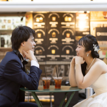 カフェテーブル越しに頬杖をついて向かい合う新郎新婦。キッチンカーのメニューボードを背にした街ロケ前撮り