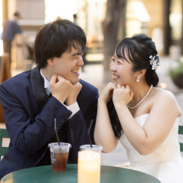 小さなカフェテーブルで頬杖をつき笑い合う新郎新婦。キャンドルの灯りが温かい街中の結婚写真