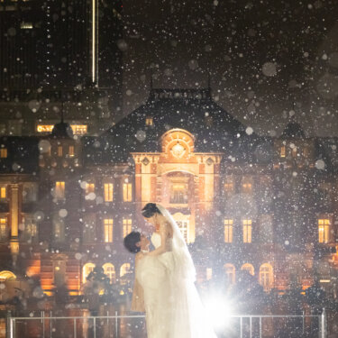 雨のベールに包まれた東京駅丸の内駅舎の前で、見つめ合いながら抱き合う新郎新婦の縦構図