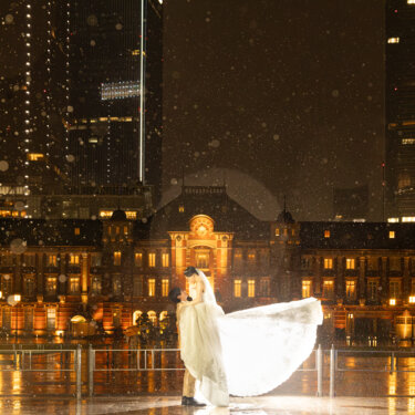 雨粒が舞う夜、ドレスの裾を大きく広げて抱き合う新郎新婦。東京駅丸の内駅舎と丸の内の高層ビルが連なる