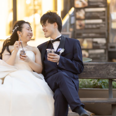 ベンチでアイスティーを手に見つめ合う新郎新婦。ネイビータキシードとパールが映える街カフェでのウェディングフォト