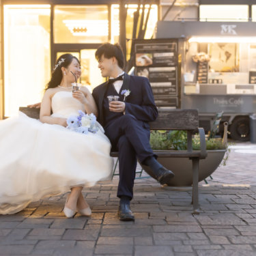 丸の内仲通りのベンチに腰かけ、ドリンク片手に笑い合う新郎新婦。キッチンカーが並ぶ街並みでの前撮り