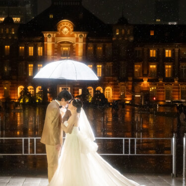 長いトレーンを広げたウェディングドレスの新婦と傘を差す新郎。東京駅丸の内駅舎のライトアップを背にした縦構図のロケーションフォト