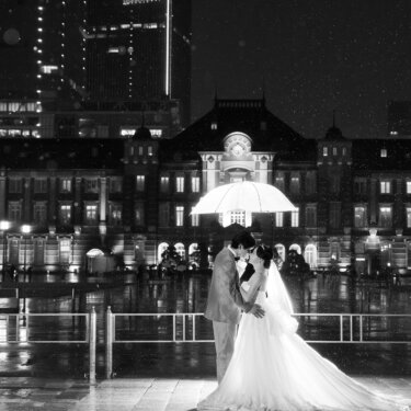 モノクロで切り取った雨の夜の東京駅前。傘の下で寄り添う新郎新婦と、丸の内駅舎・高層ビルのシルエット
