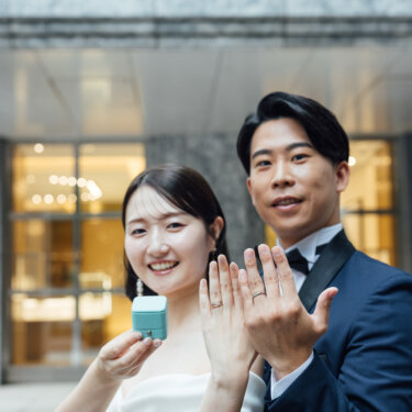 リングボックスを手に、そろって左手の指輪を見せて微笑むネイビータキシードの新郎とドレスの花嫁の結婚写真