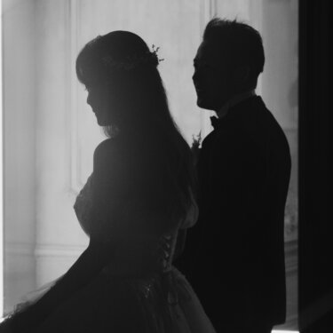 窓明かりに浮かぶ新郎新婦の横顔のシルエット。編み上げのウェディングドレスと黒タキシードのモノクロポートレート