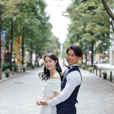 並木道の真ん中で花嫁を後ろから抱き寄せてカメラに微笑む新郎。丸の内の街並みで撮る自然体のウェディングフォト