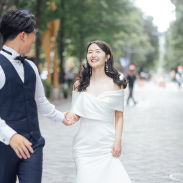 丸の内の並木道を手をつないで小走りし、振り返って笑い合う新郎新婦。躍動感のある街歩きのロケーションフォト