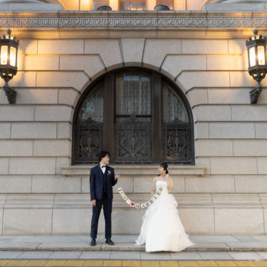 アーチ扉とランタンが並ぶ石造建築の前で'JUST MARRIED'のガーランドを広げる新郎新婦。丸の内での洋装前撮り