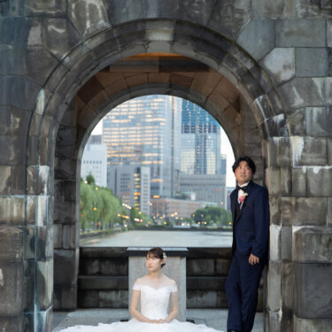 石のアーチの下でドレスを広げて座る花嫁と、柱にもたれる新郎。アーチの向こうに丸の内のビル群が見える構図の前撮り