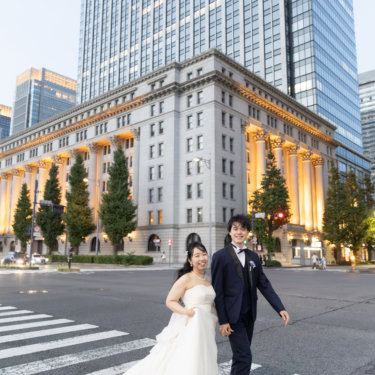 重厚な列柱の歴史的建築を背に、横断歩道を並んで渡る新郎新婦。夕暮れの丸の内で撮る洋装ロケーション前撮り