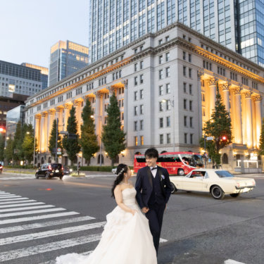 重厚な列柱の建物とクラシックカーを背に、横断歩道を渡る新郎新婦。夕暮れの丸の内で撮る洋装ロケーション前撮り