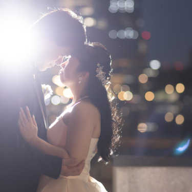 強い逆光の中で新郎に寄り添うウェディングドレスの花嫁と、奥に広がる東京駅周辺の夜景のボケ。東京駅で撮るナイトフォト前撮り