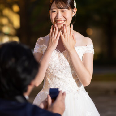 指輪を差し出す新郎の前で、両手を頬に添えてはじける笑顔を見せる花嫁。夜の街で撮るプロポーズシーンの前撮り
