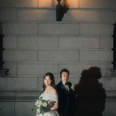 石積みの壁に灯る外灯の下、背中合わせに立つ新郎新婦。オフショルダーのウェディングドレスと黒タキシードで撮る夜のフォトウェディング