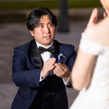 ひざまずいて指輪を差し出す新郎と、口元に手を寄せて喜ぶ花嫁を肩越しに写したプロポーズ前撮り