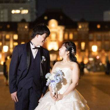 ライトアップされた東京駅丸の内駅舎の前で見つめ合う新郎新婦。ブルーのブーケとネイビータキシードのナイトフォト