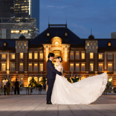 ブルーアワーの空と赤レンガの東京駅駅舎の前で、ドレスの裾を大きくなびかせる花嫁と新郎のロケーションフォト