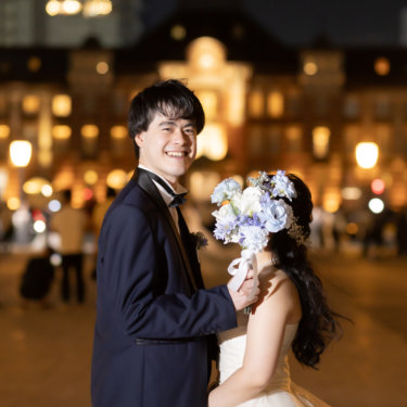 ブルーのブーケで顔を隠す花嫁と、振り返って笑う新郎。東京駅丸の内駅舎の灯りを背にした夜の結婚写真