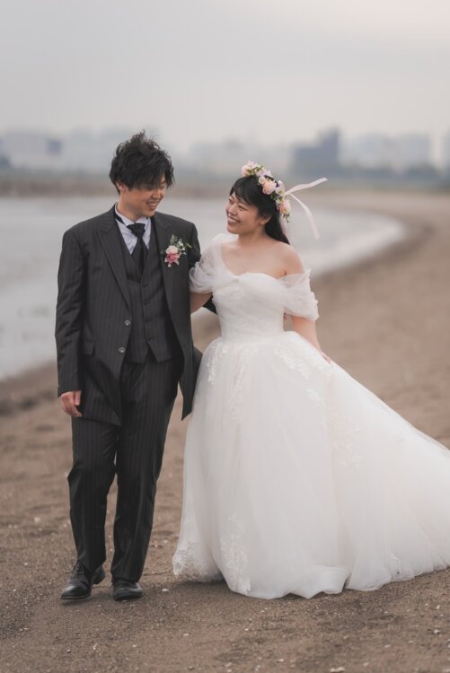 海辺の砂浜を笑顔で並んで歩く、ストライプのブラックタキシードの新郎と花冠の花嫁。大きなサイズでも着こなせるタキシードで叶える結婚写真（ブラックラージ／衣装番号TP08）