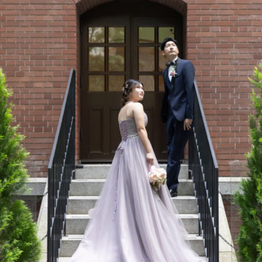赤レンガの洋館の階段に並ぶ新郎新婦。くすみパープルのカラードレスの裾が階段に流れる縦構図の結婚写真