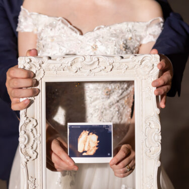 白い額縁越しにエコー写真を見せる新郎新婦の手元のアップ。家族が増える報告にもぴったりなマタニティのウェディングフォト