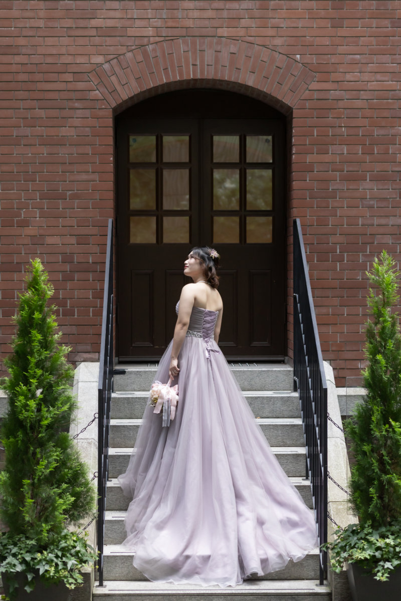 赤レンガの洋館の階段でドレスの裾を大きく広げ、振り返る新婦。くすみパープルのカラードレスで撮る丸の内の前撮り