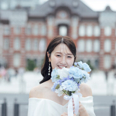 ぼかした東京駅丸の内駅舎を背景に、ブルーの紫陽花のブーケで口元を隠して微笑むオフショルダードレスの花嫁の前撮り写真