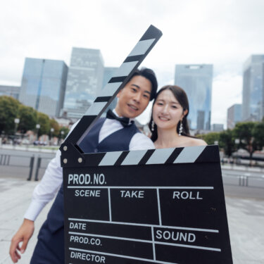 丸の内の高層ビルを背景に、カチンコを手前に差し出して笑う新郎新婦。映画のワンシーンのような遊び心あるフォトウェディング