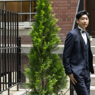 赤レンガの洋館の階段に立つ新郎のソロカット。ネイビーのタキシードとピンクのブートニアが緑に映える