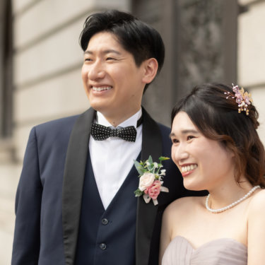 笑顔でこちらを見上げる新郎新婦のバストアップ。ネイビーのタキシードとパールのネックレス、ヘッドドレスが上品な結婚写真