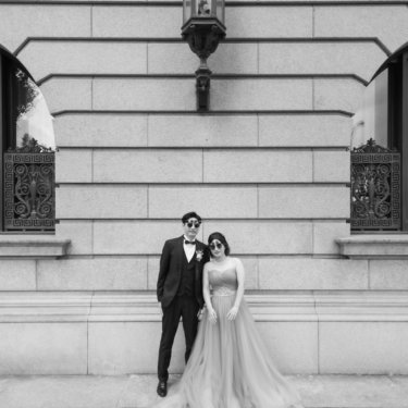 サングラス姿でポーズを決める新郎新婦のモノクロ写真。ガス灯と鉄の装飾窓が並ぶ石造り建築を背にしたおしゃれな一枚