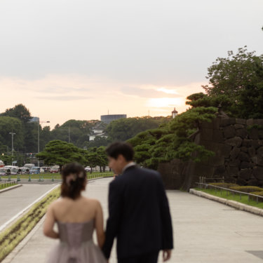 沈む夕日に向かって歩く新郎新婦の後ろ姿。松の木と石垣、遠くに望む櫓を入れた夕景のフォトウェディング