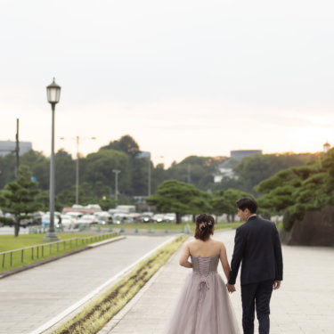編み上げのカラードレスの新婦と新郎が手をつないで歩く後ろ姿。松と石垣が続く夕暮れの散策路での前撮り