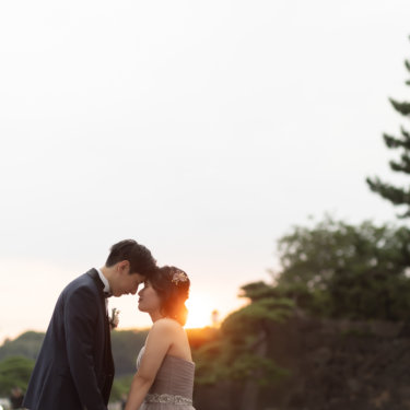 夕日に照らされながら額を寄せ合い、手をつなぐ新郎新婦。石垣と松並木が続く夕暮れの散策路でのロケーションフォト