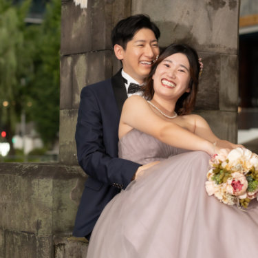 石造りの門にもたれて声を上げて笑う新郎新婦。くすみピンクのカラードレスとブーケが映える自然体の前撮り