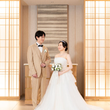 白バラのブーケを持つティアラ姿の新婦と、ベージュのスリーピースタキシードの新郎が並ぶ全身カット。組子格子の和モダンチャペルでの洋装前撮り