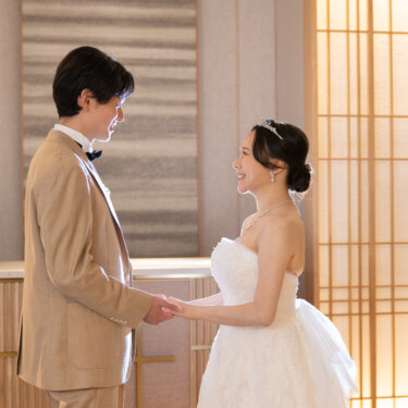 ベージュのタキシードの新郎と白いウェディングドレスの新婦が向かい合って手を取り、笑顔を交わす。障子の光がやわらかい和モダンチャペルでの洋装前撮り