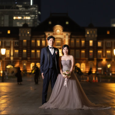 ライトアップされた東京駅丸の内駅舎を背に並ぶ新郎新婦の全身。くすみピンクのカラードレスとネイビーのタキシードで撮る夜のフォトウェディング