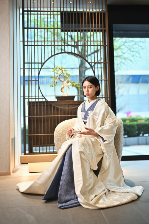 白地の打掛に紺の掛下を合わせた花嫁が、丸窓の衝立と盆栽を背にソファへ腰かける。和モダンなスタジオでの和装前撮り