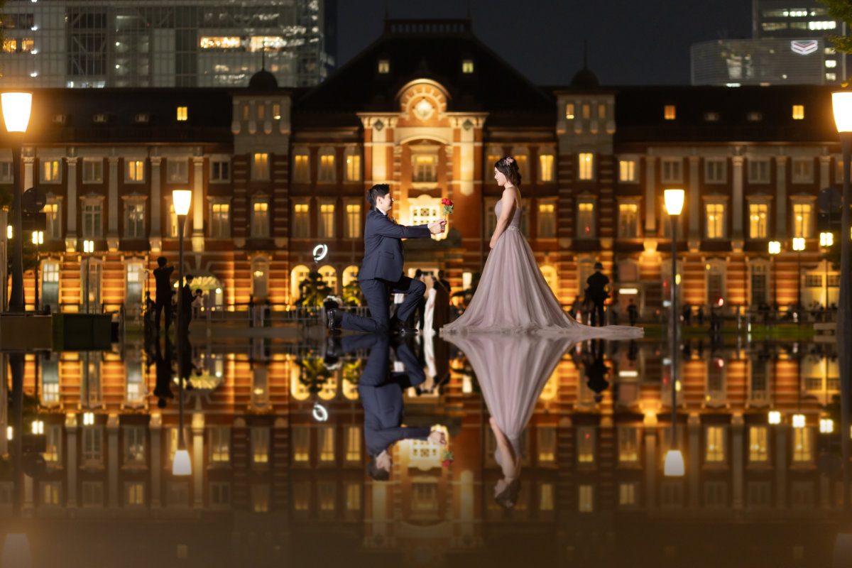 水鏡のリフレクション撮影。東京駅丸の内駅舎の前で片膝をつき、赤いバラを差し出す新郎とカラードレスの新婦