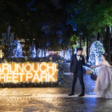 MARUNOUCHI STREET PARK のネオンサインとイルミネーションを背景に、手をつないで歩く新郎新婦。丸の内仲通りの夜の前撮り