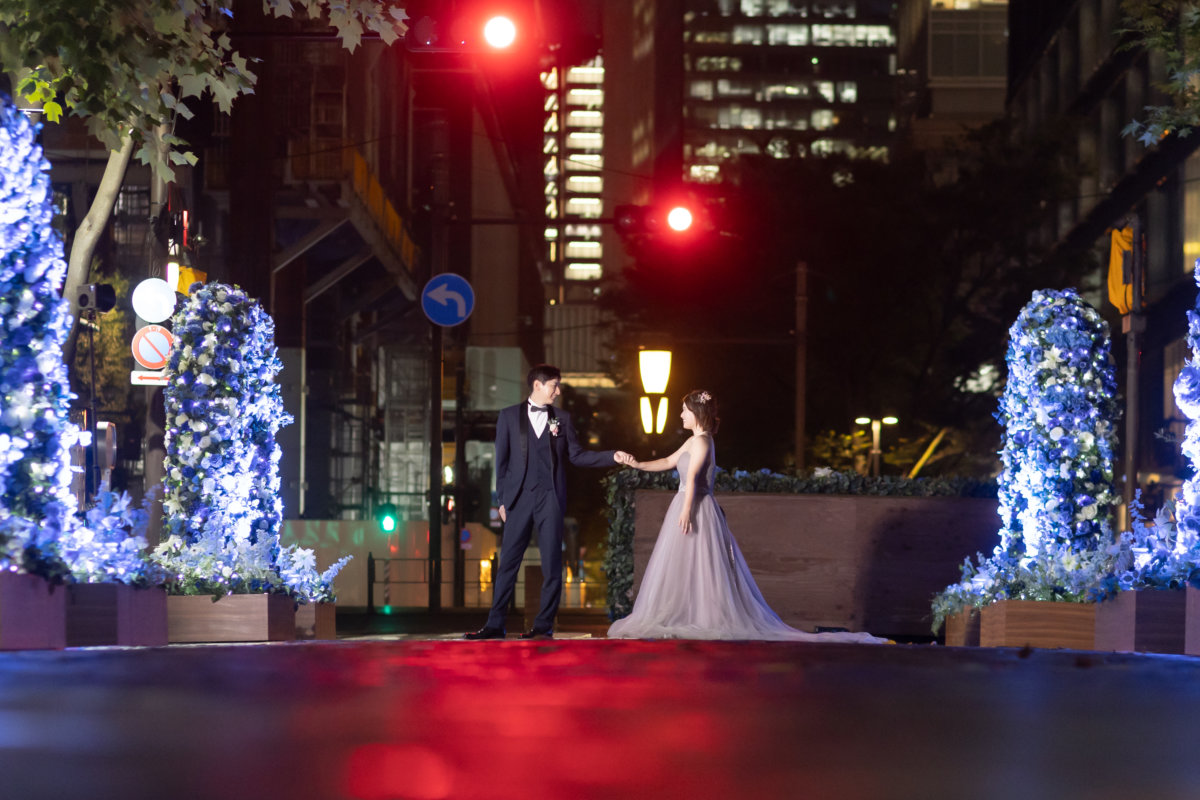青いイルミネーションが灯る丸の内仲通りで手を取り合う新郎新婦。くすみピンクのカラードレスで撮る夜のロケーションフォト