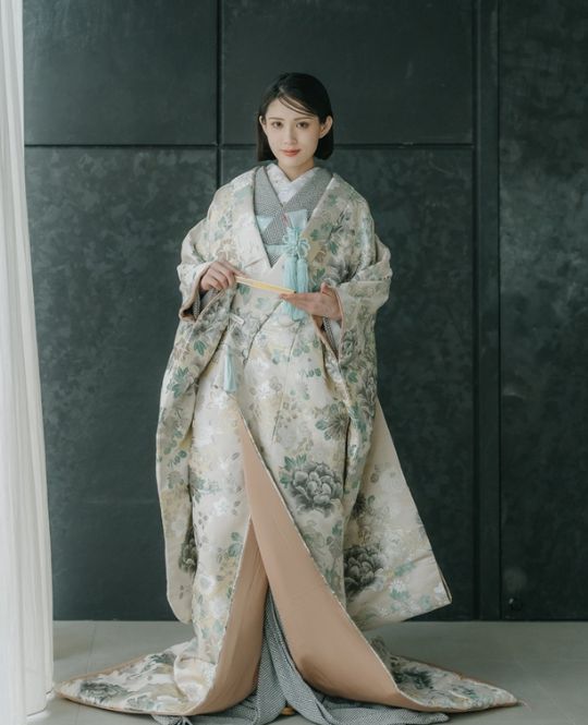 黒い背景の前で、扇子を持ち佇む花嫁、緑がかった刺繍の掛下姿。