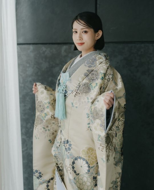 灰色の背景の前で、扇子を持ち佇む花嫁、青みがかった刺繍柄の白い掛下姿。