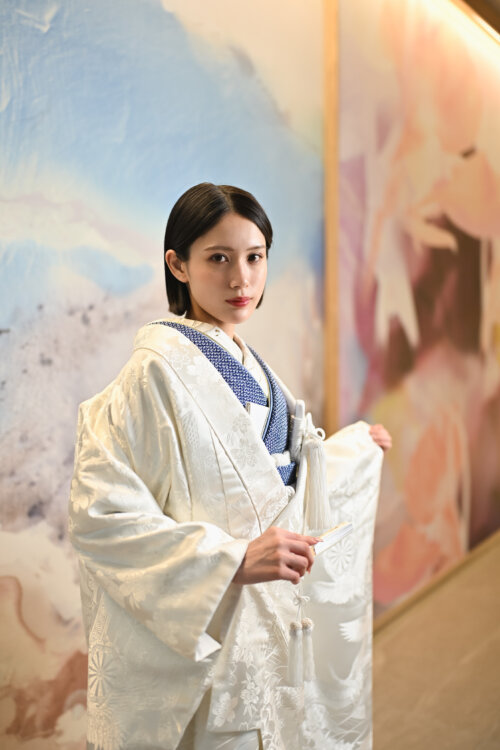 白地の打掛に紺の掛下を合わせ、末広を手にした花嫁。淡い色彩のアートウォールを背にした和装前撮りのポートレート