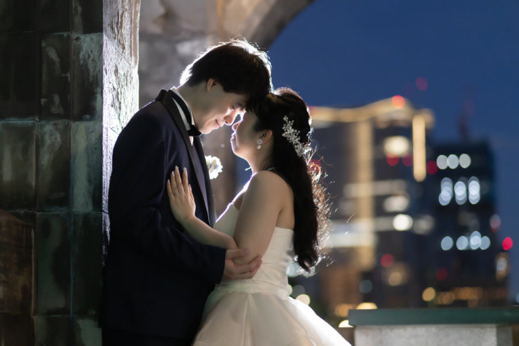 石造りの門の陰で額を寄せ合う新郎新婦。夜景のボケが背景に広がる、ネイビータキシードとウェディングドレスの結婚写真