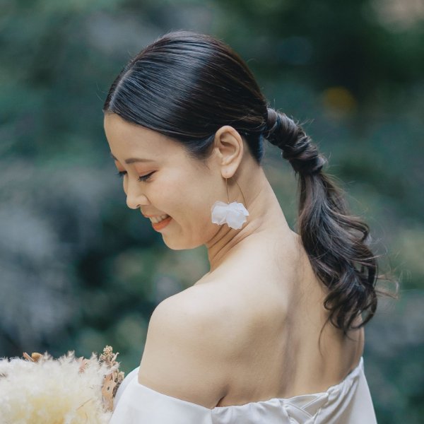 白い花のイヤリングをつけ、緑を背景に微笑みながらうつむく新婦の後ろ姿ヘアスタイル。