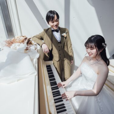 窓辺の白いピアノを弾くウェディングドレスの花嫁と見守る新郎。南青山のスタジオで撮る洋装前撮り。