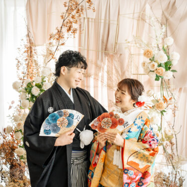 花のアーチの前で『夫』『嫁』の扇子を手に見つめ合う、黒紋付袴の新郎と色打掛の花嫁の和装スタジオフォト